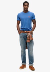 Camiseta de algodón azul con un pequeño gráfico rojo, combinada con jeans de mezclilla desgastados y una camisa de cuadros atada a la cintura. Zapatos de cordones marrones.