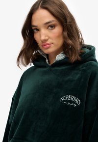 Gros pull en côtes vert avec une poche kangourou et un logo "SUPERDRY" brodé en blanc. Tissu texturé et grande capuche drapée.