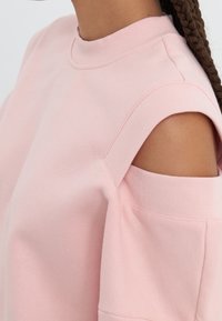 Light pink sweatshirt with a high neck, cut-out shoulder design, and textured fabric. Features a relaxed fit and minimal stitching details.