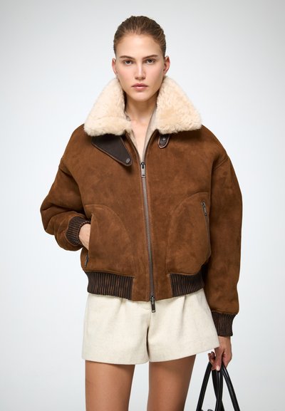 Giacca bomber in suede marrone con colletto in pelliccia crema, polsini a coste e due tasche con zip. Dettaglio della zip argentata sul davanti.
