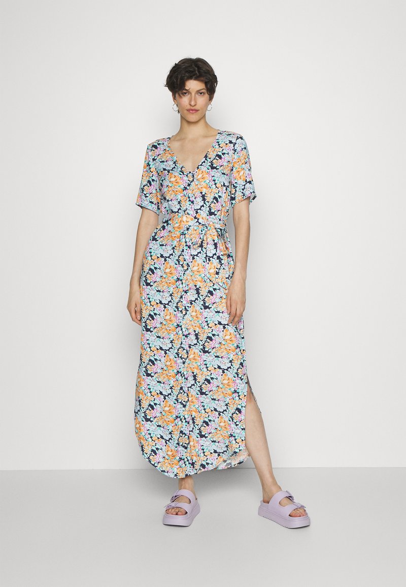 Vestido maxi floral com decote em V, mangas curtas, flores azuis, laranjas e rosas sobre um fundo escuro; cintura ajustada e fendas laterais. Calçado roxo.