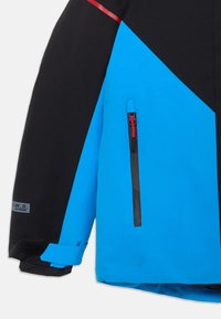 Une veste bleue et noire avec un design géométrique, comprenant une poche zippée avec des accents rouges et un détail de poignet en Velcro.