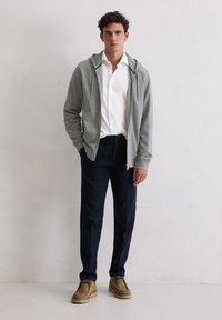 Felpa grigia con zip sopra una camicia bianca con bottoni, abbinata a pantaloni navy e scarpe in suede marrone chiaro. Tessuto strutturato e vestibilità casual.