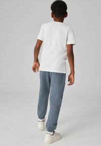 Bílá polokošile s krátkým rukávem a šedé tepláky s elastickými manžetami, které mají kapsy a decentní branding. Bílé tenisky doplňují celý outfit.