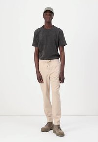 T-shirt gris à manches courtes avec le logo "GUESS", pantalon beige à cordon, et bottes à lacets couleur sable. Design simple avec une coupe décontractée.