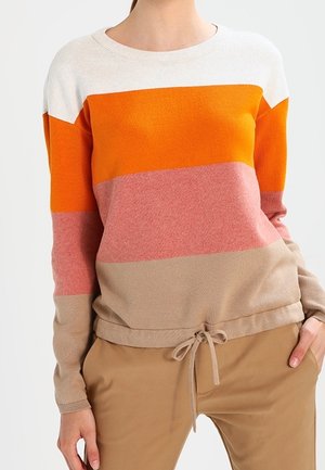 Femme portant un pull rayé blanc, orange, rose et beige avec un cordon de serrage à l’ourlet, associé à un pantalon marron clair.