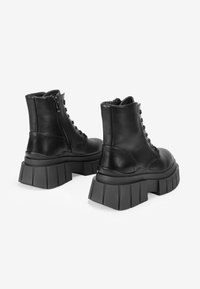 Stivali neri in pelle con lacci alla caviglia, dotati di suole a piattaforma spesse e scanalate, presentati di spalle e leggermente inclinati.