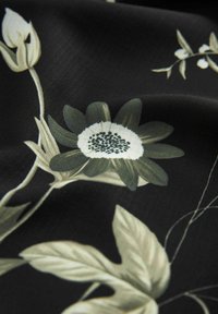 Schwarzer Stoff mit einem floralen Muster aus grünen Blättern und weißen Blumen. Die Textur wirkt glatt und das Design ist filigran.