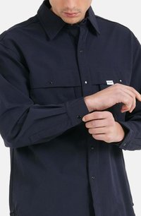Camicia blu navy realizzata in tessuto morbido, con colletto, chiusura anteriore con bottoni e polsini regolabili con dettagli in hardware nero.