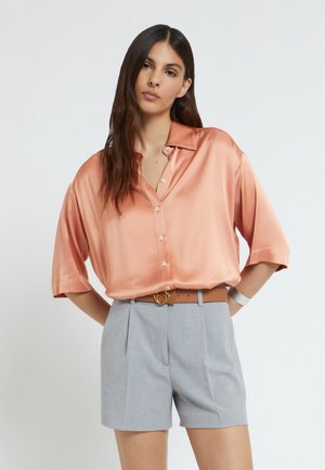 Vrouw met lang donker haar die een losse perzikkleurige satijnen blouse draagt, ingestopt in lichtgrijze, op maat gemaakte korte broek met een bruine riem, staand met handen achter de rug.