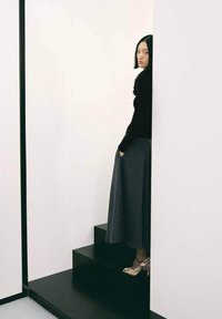 Modella che indossa un top nero a maniche lunghe e una gonna lunga grigia scura, in piedi su scale nere contro uno sfondo bianco.