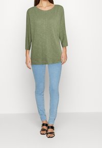 Olivgrünes Langarmoberteil mit weitem Ausschnitt und weicher Textur, kombiniert mit hellblauen Skinny Jeans und schwarzen Riemchensandalen.