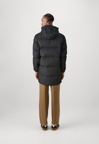 Zwarte pufferjack met een capuchon, voorzien van gequiltte delen en een middellange snit. Dragen over beige broek en zwarte schoenen.