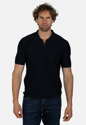 Hombre con pelo rizado que lleva un polo negro de manga corta y vaqueros azul oscuro, de pie frente a un fondo claro y liso.