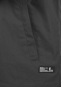 Musta kangas, jossa on sileä pinta, sisältää läppätaskun ja tunnisteen, jossa lukee "SOLID EST. 1999 DENMARK" brändin yksityiskohtineen.