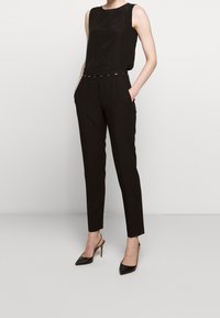 Černý top bez rukávů spárovaný s černými kalhotami na míru s úzkým střihem, předními kapsami a lesklými černými lodičkami na vysokém podpatku.