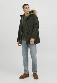 Dark green parka jacket with a fur-lined hood, silver snap closures, two front pockets, worn over a patterned green sweater and blue jeans.