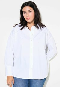 Camicia bianca a maniche lunghe con bottoni e colletto, presenta sottili decorazioni sulle spalle e una vestibilità rilassata, indossata con jeans blu.