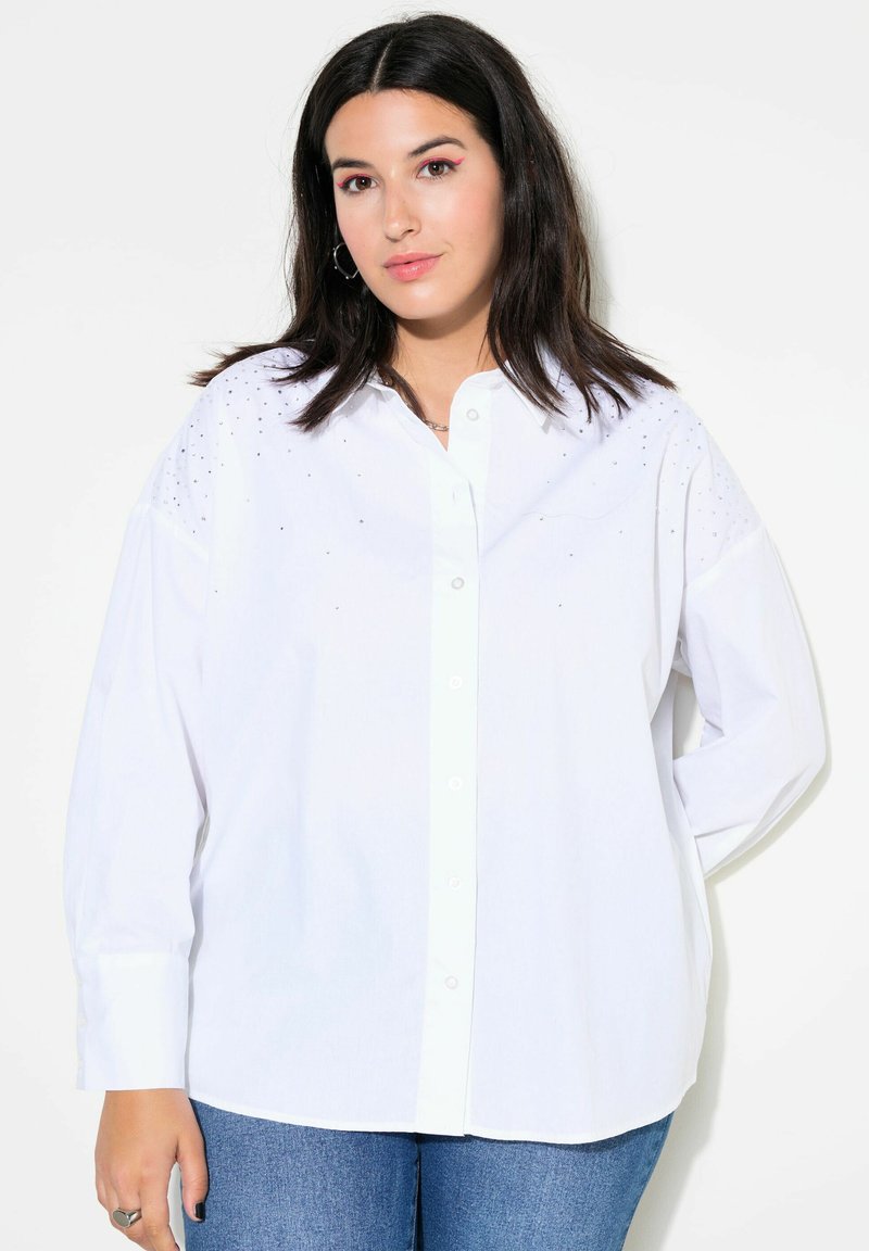 Camicia bianca a maniche lunghe con bottoni e colletto, presenta sottili decorazioni sulle spalle e una vestibilità rilassata, indossata con jeans blu.