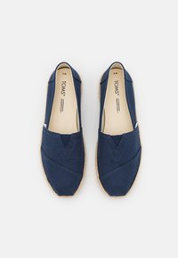 Een paar marineblauwe TOMS instap schoenen met beige binnenzolen op een witte achtergrond, maat 9M, met jute-wikkelsols.