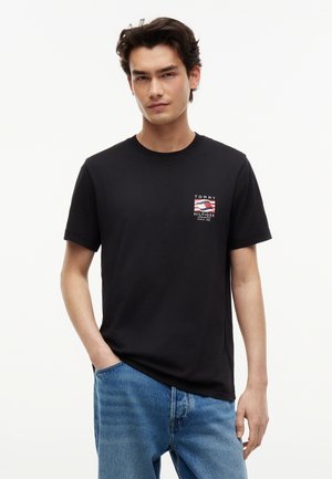 Νεαρός άνδρας με μαύρο t-shirt Tommy Hilfiger με λογότυπο στο στήθος, μπλε τζιν, στέκεται με το ένα χέρι στην τσέπη μπροστά σε λευκό φόντο.