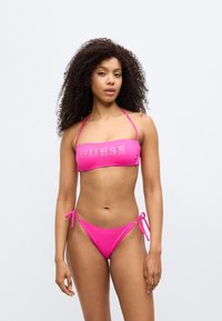 Σετ ροζ μπικίνι που αποτελείται από τοπ μπαντάνα με τη λέξη "GUESS" από στροντίνια και εσώρουχα με ρυθμιζόμενους δεσίμους στο πλάι. Λείο, ελα στικό ύφασμα.