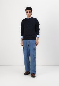 Felpa blu navy con logo Tommy Hilfiger in rilievo, abbinata a ampi jeans cargo blu con tasche e scarpe marroni.