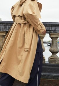 Trench-coat beige avec capuche, ceinture décorative et design en couches. Accentué par une finition texturée, porté sur un pantalon marine avec des rayures blanches.