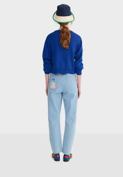 Personne aux longs cheveux roux attachés en queue de cheval portant un bob beige et bleu marine, un pull bleu, un jean bleu clair avec un patch coloré, et des chaussures multicolores.