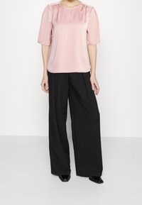Blusa di raso rosa chiaro con maniche a sbuffo e scollatura arricciata, abbinata a pantaloni neri larghi e scarpe nere a punta.