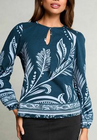 Blusa de manga larga en color teal que presenta un escote en forma de ojo de cerradura, estampados botánicos en blanco, puños elasticados y un dobladillo estampado. Textura de tela suave.
