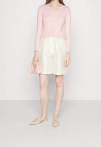 Mujer que lleva un cárdigan abotonado en rosa pálido, pantalones cortos de satén crema, zapatos de lona beige con estampado y sostiene un pequeño clutch recogido rosa.