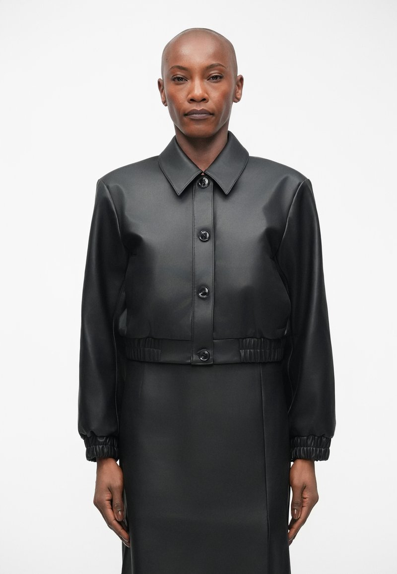 Crna skraćena jakna od sjajnog materijala, s prednjim kopčanjem na gumbe, raširenim ovratnikom i elastičnim manžetama za prilagođeni izgled.