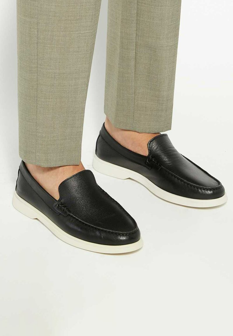 Black leather slip-on shoes with a textured finish, featuring white rubber soles and decorative stitching along the edges.