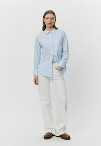 Cămașă butonată bleu deschis, cu un buzunar, asortată cu pantaloni albi și pantofi maro. Țesătura pare netedă și ușoară.