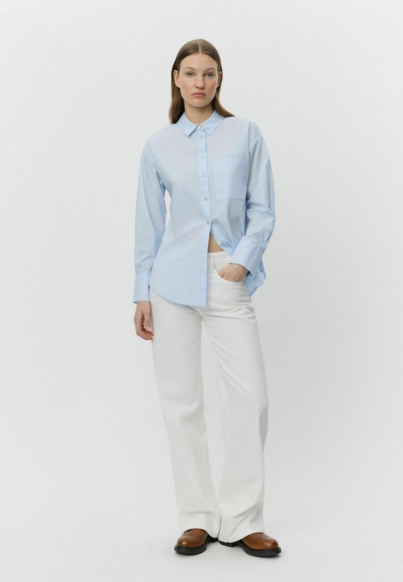 Cămașă butonată bleu deschis, cu un buzunar, asortată cu pantaloni albi și pantofi maro. Țesătura pare netedă și ușoară.