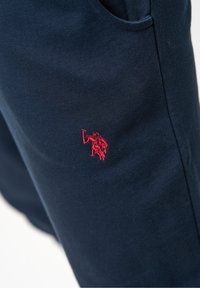 Tmavě modré bavlněné tepláky s malým červeným vyšitým logem. Materiál je měkký s hladkou texturou a bočními kapsami.