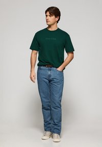 Mörkgrön bomullströja med texten "Polo Club", i kombination med ljusblå denimjeans och beige sneakers. Enkel, avslappnad passform.