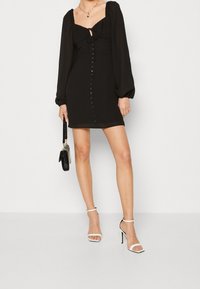 Svart miniklänning med puffärmar, fyrkantig halsringning, knytfront och knappdetaljer. Bärs tillsammans med vita remsandaler och en svart handväska.