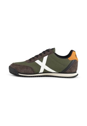 Zapatilla verde y marrón con cordones negros, diseño lateral blanco en forma de X, suela negra y lengüeta naranja en el talón sobre fondo blanco.