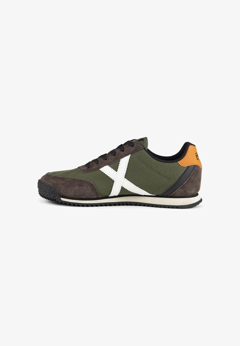 Baskets vertes et marron avec lacets noirs, motif X blanc sur le côté, semelle noire et languette orange au talon sur fond blanc.