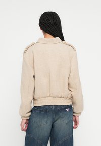 Casaco bege texturizado com gola alta, bainha canelada e ombreiras com ferragens douradas. Combina com jeans azul escuro.