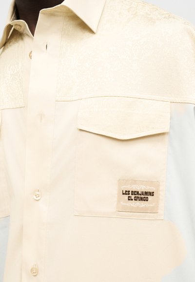 Chemise beige à boutons avec une poche à rabat sur la poitrine portant une étiquette "Les Benjamins El Gringo".