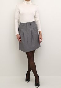 Falda gris plisada con cintura ajustada y bolsillos laterales, combinada con un suéter de cuello alto de manga larga blanco y zapatos de tacón alto negros y brillantes.