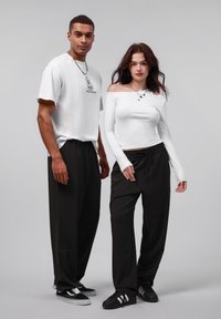 Chemise blanche à manches courtes avec un graphique, pantalons rayés noirs et baskets noires. Le modèle à gauche a une chaîne en argent ; le modèle à droite porte un haut à manches longues.