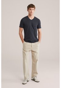 Jaunas vyras stovintis kišenėse rankomis, dėvintis juodą V formos kaklo marškinėlį, smėlio spalvos kelnes ir baltus sportinius batelius su žaliomis juostelėmis.