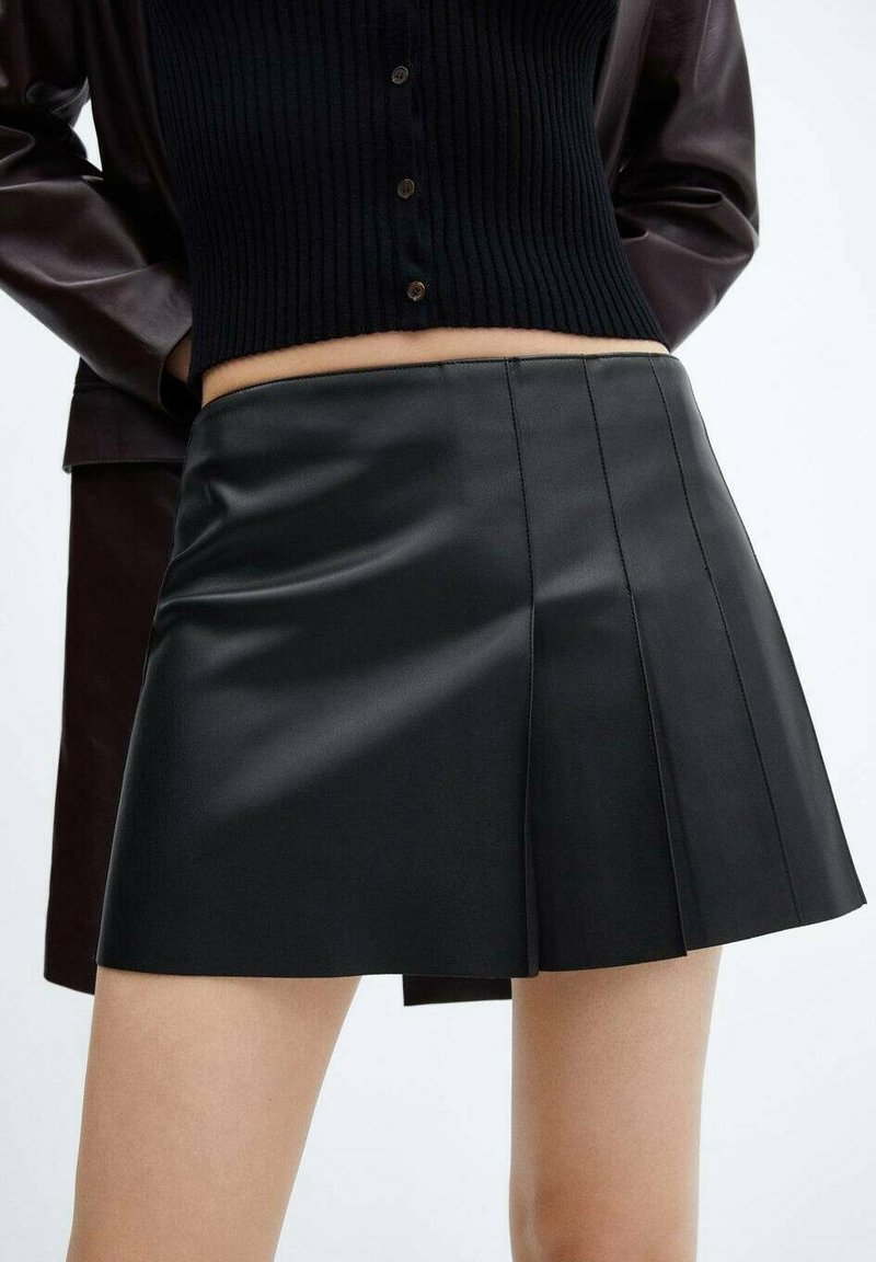 Mini-jupe noire plissée en cuir portée avec un haut court noir côtelé à boutons et une veste extérieure sombre sur fond blanc.