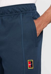 Tamsiai mėlynos sportinės kelnės su elastine juosmens juosta, šonine kišene ir daugiaspalviu stačiakampio formos Nike logotipo lopu ant šlaunies.