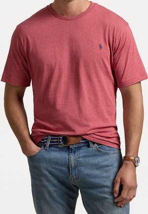 Mann trägt ein rotes T-Shirt mit kurzen Ärmeln und einem kleinen blauen Logo, einen blau gestreiften Gürtel, Jeans und eine braune Lederuhr am linken Handgelenk.