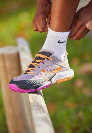 Ruce upravují bílé ponožky Nike na noze nosící šedé a oranžové běžecké boty Nike, která stojí na dřevěném plotu venku.